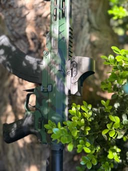 Close-up of the AR-15 rifle with camouflage pattern leaning against a tree on its right side.