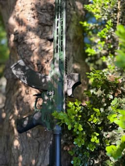 AR-15 rifle with camouflage pattern leaning against a tree on its right side.