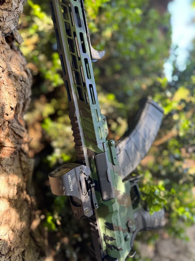 AR-15 rifle with camouflage pattern leaning against a tree on its left side.